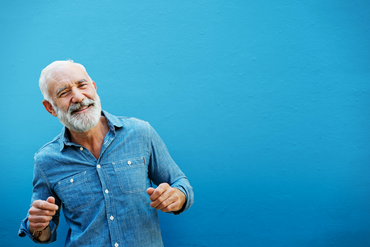 Kék háttér előtt kék inges, ősz, szakállas férfi táncoló mozdulatot tesz