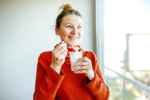 piros pulóveres idősebb nő mosolyogva joghurtot eszik