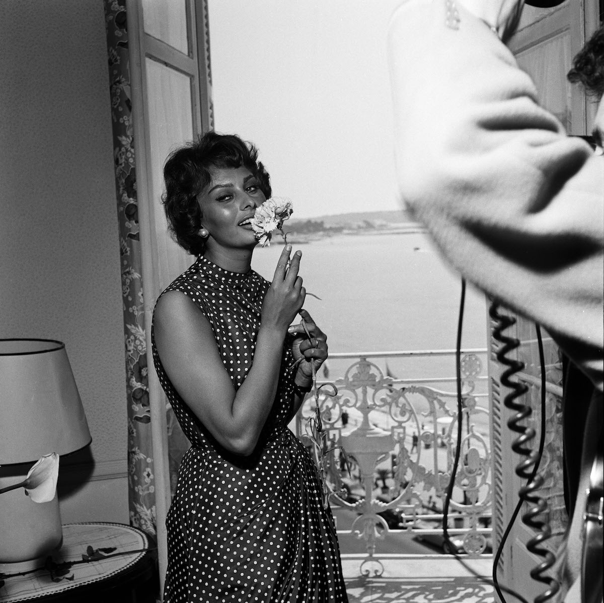 Sophia Loren virágot szagol és mosolyog, a háttérben egy erkély és a tenger Cannes-ban