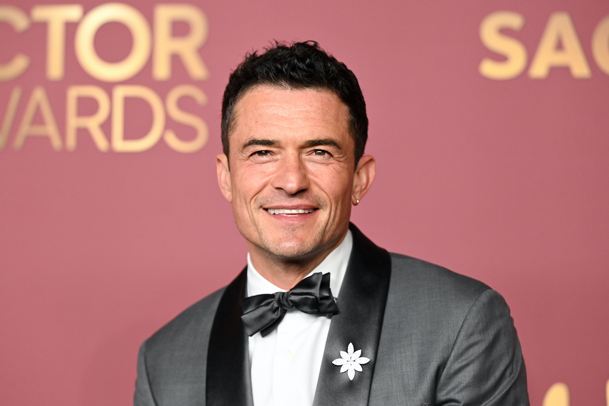 Orlando Bloom rózsaszín háttér előtt csokornyakkendőben nevet