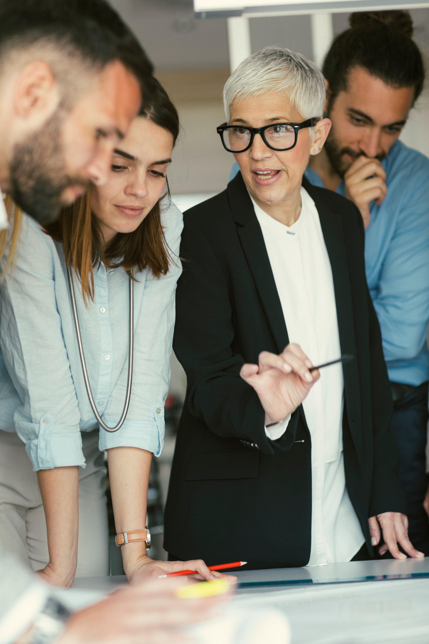 narcisztikus-kollega-vezetoi-pozicio | Marie Claire Megbeszélés egy irodában, nők és férfiak figyelnek egy idősebb, szemüveges nőt, aki magyaráz