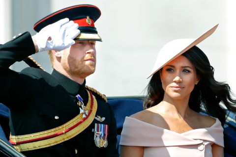 Harry herceg szalutál, mellette Meghan Markle fehér kalapban és ruhában néz rá