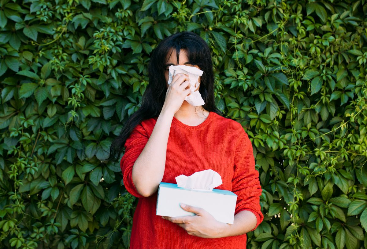 Allergiás nő áll zöld növényfal előtt piros pulcsiban, papírzsebkendőt tartva az orra elé
