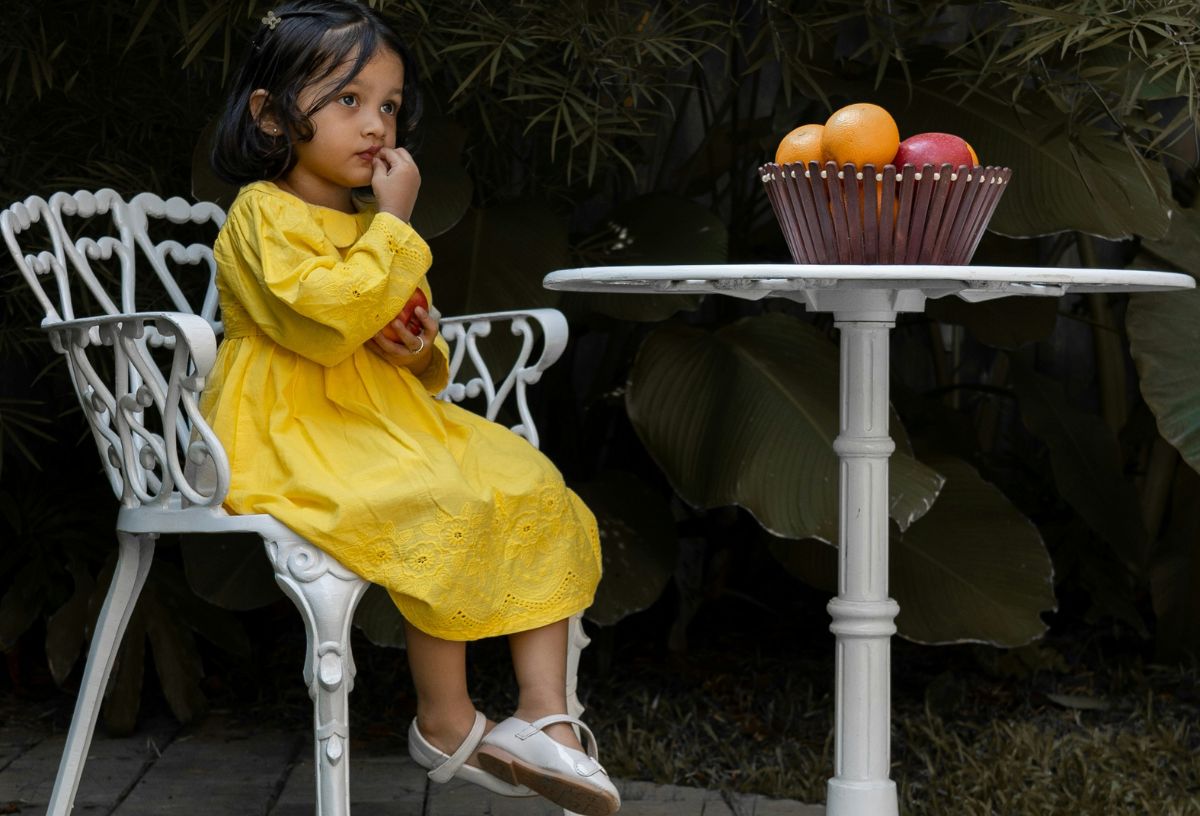 Barna bőrű, fekete hajú kislány sárga ruhában fehér kerti széken ül, mellette kör asztal gyümölcskosárral