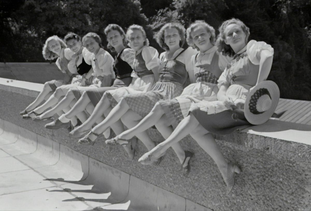 Régi fekete-fehér képen nők ülnek kőfalon sorban ugyanabban az irányban keresztbetett lábbal