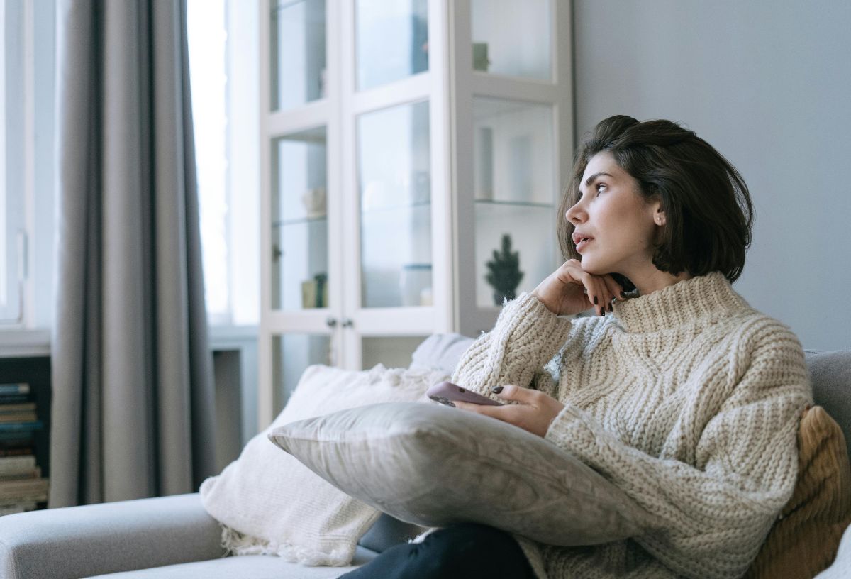 5 tipp, hogyan hozd helyre testbeszéddel a kapcsolatodat vita után | Marie Claire Barna bob hajú nő kötött pulcsiban ül a kanapén, gondolkodva néz, ölében díszpárna, kezében telefon