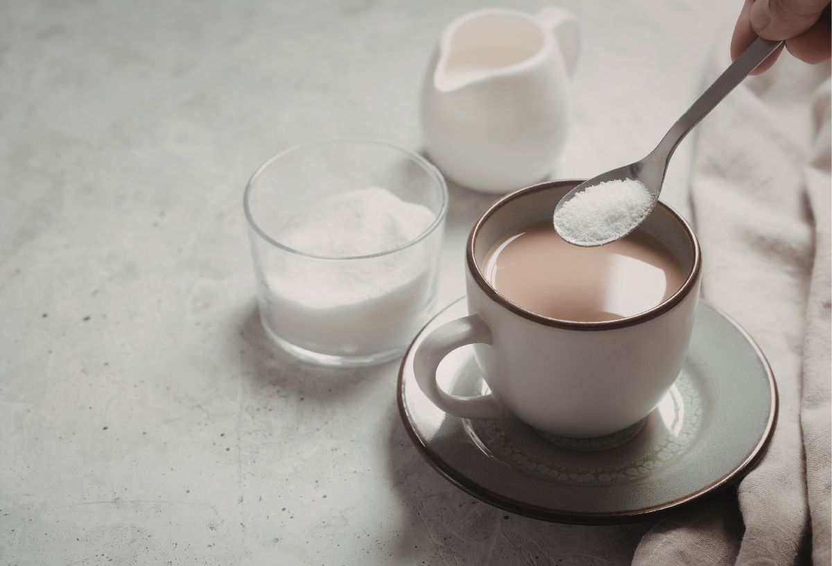 Kávéscsészébe kiskanállal szukralózt ad cukortartóból kéz, mellette fehér tejkiöntő