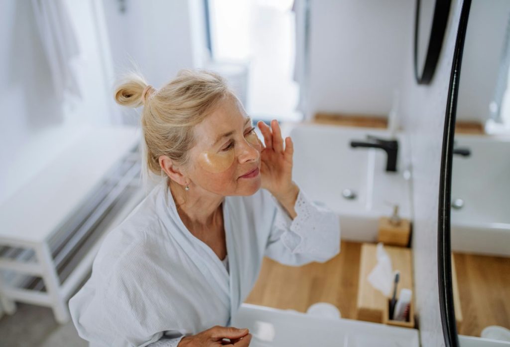 Idősebb nő tükörben nézi a bőrét, szeme alatt ceramidos maszk