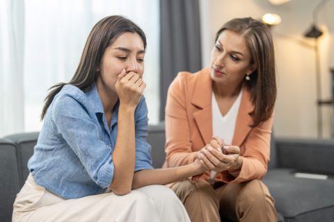Két fiatal nő ül a kanapén, az egyik eltakarja a száját és maga alatt van, míg a másik fogja a kezét és vigasztalja