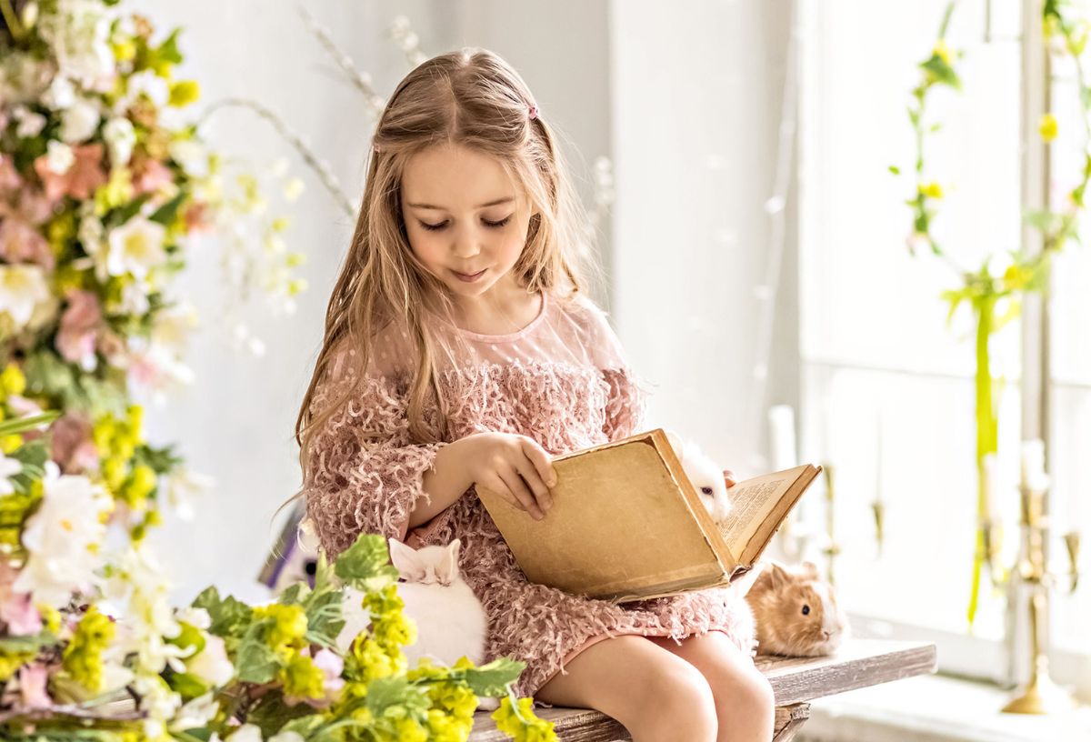 Szőke, hosszú hajú kislány csinos ruhában húsvéti nyulak, sárga virágok között ülve könyvet olvas