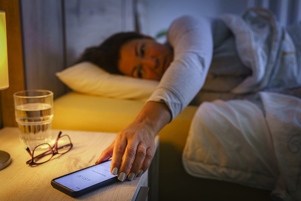 Egy nő az ágyban, mobiltelefon után nyúl az éjjeliszekrényen