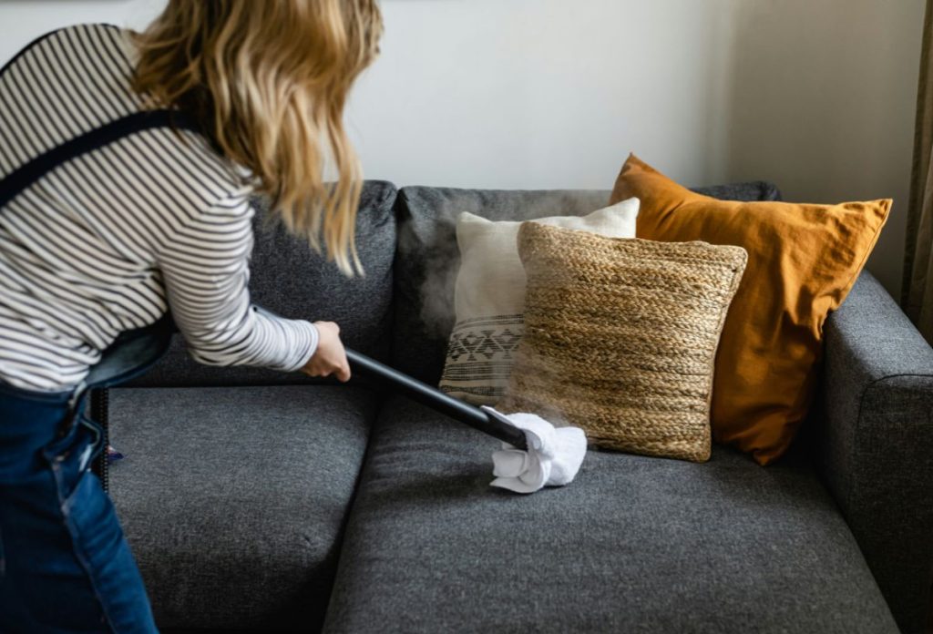 Nő háttal állva gőzölős takarítógéppel tisztítja a szürke kanapéhuzatot