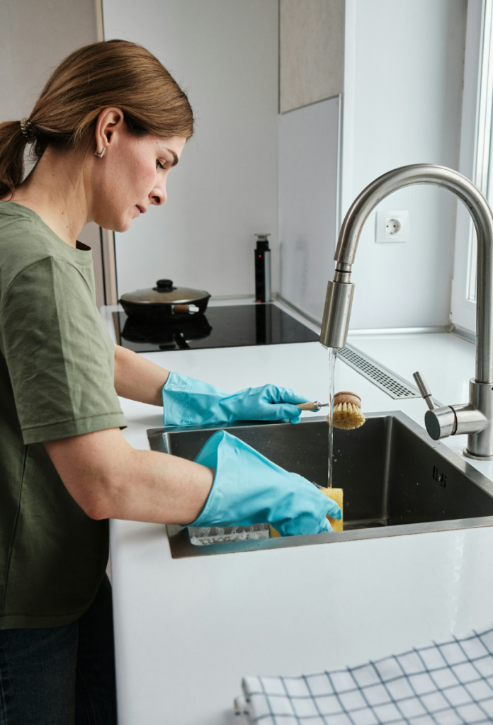 Nő kék gumikesztyűben a mosogató felett állva kimossa a szivacsot, másik kezében súrolókefe