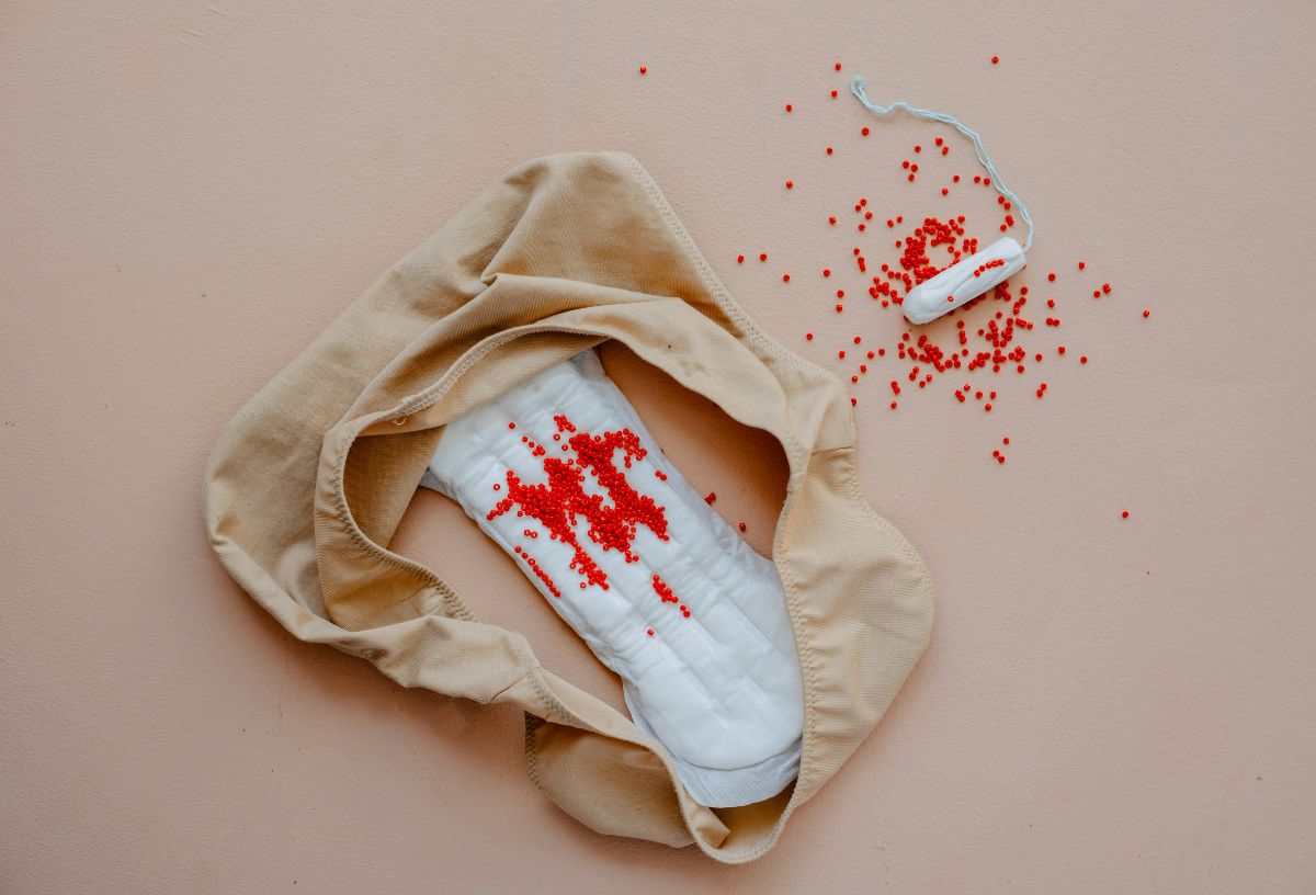 Bézs háttéren testszínű bugyi egészségügyi betéttel, mellette tampon, piros flitterek a menstruációs vért szimbolizálják
