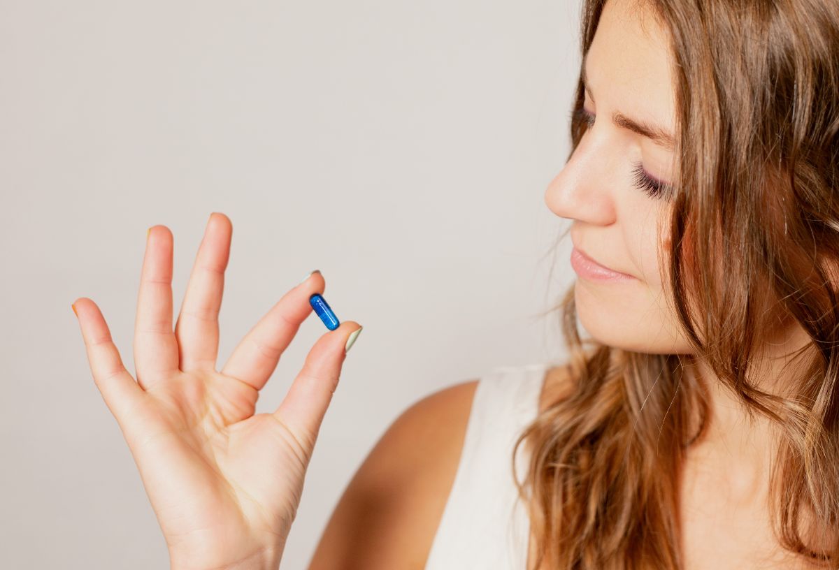 Nő két ujja között kék kapszulát tart és ránéz