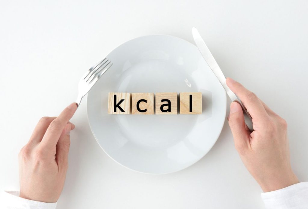 Fehér tányéron kcal felirat fakockákon betűnként, kéz evőeszközt tart