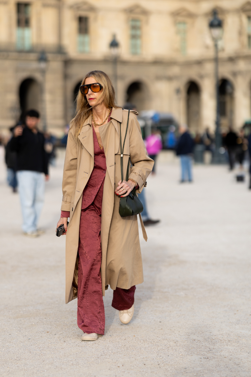 Ilyen egy stílusos vendég a Schiaparelli 2026-os őszi-téli bemutató előtt: a rózsaszín overall és a bézs trench coat párosítása telitalálat, amit olívazöld crossbody táskával, napszemüveggel és kényelmes, bézs balerinacipővel tett teljessé.