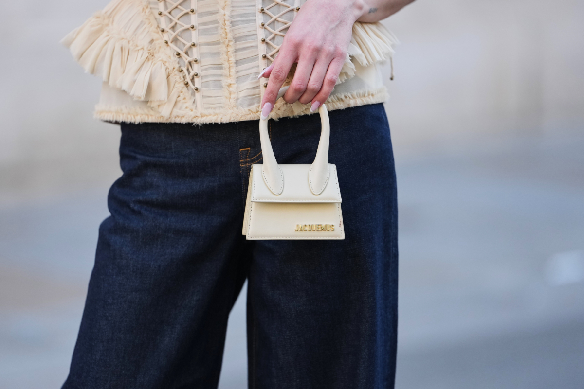Egy vendég bézs színű, fodros tüllből készült, ejtett vállú csipkefűzőt és sötétkék, széles szárú farmernadrágot visel, amelyet egy bézs színű Jacquemus bőr mikrotáskával egészített ki. A fotó a Zimmermann bemutatója előtt készült a párizsi divathéten