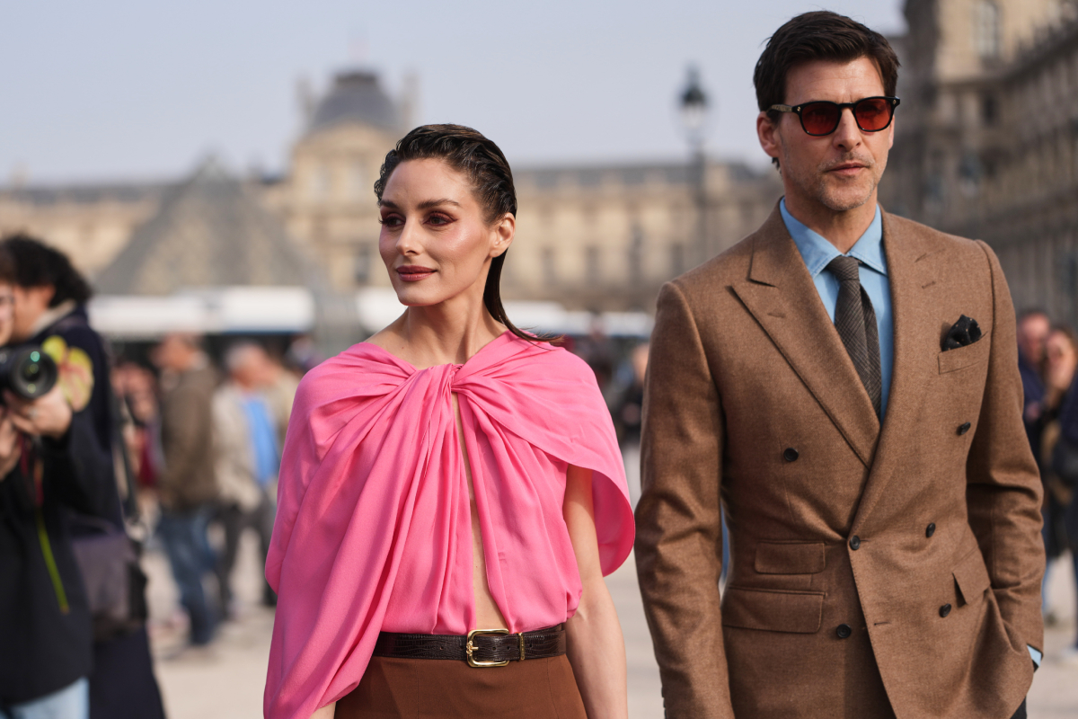 Olivia Palermo (balra) sötétbarna hajjal, egy élénkrózsaszín, elöl csomózott drapériás szaténblúzt és egy középbarna, magas derekú ceruzaszoknyát visel, fekete lakkbőr övvel kiegészítve. Johannes Huebl (jobbra) sötétbarna, szögletes napszemüvegben, kék ingben, fekete nyakkendőben és barna zakóban látható az Elie Saab bemutatója előtt a párizsi divathéten