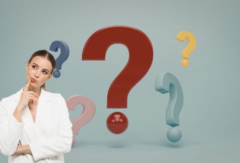 nő fehér blézerben gondolkodva dönti oldalra a fejét, háttérben zöldesszürke szín, öt színes kérdőjel