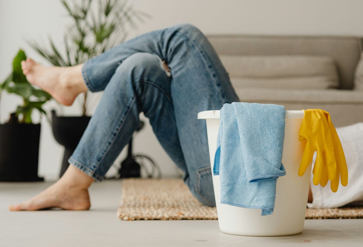 Nő tavaszi nagytakarítás közben pihenésképpen lefeküdt a padlóra, lábait keresztbe rakta, előtérben fehér vödör gumikesztyűvel és kék törlőronggyal