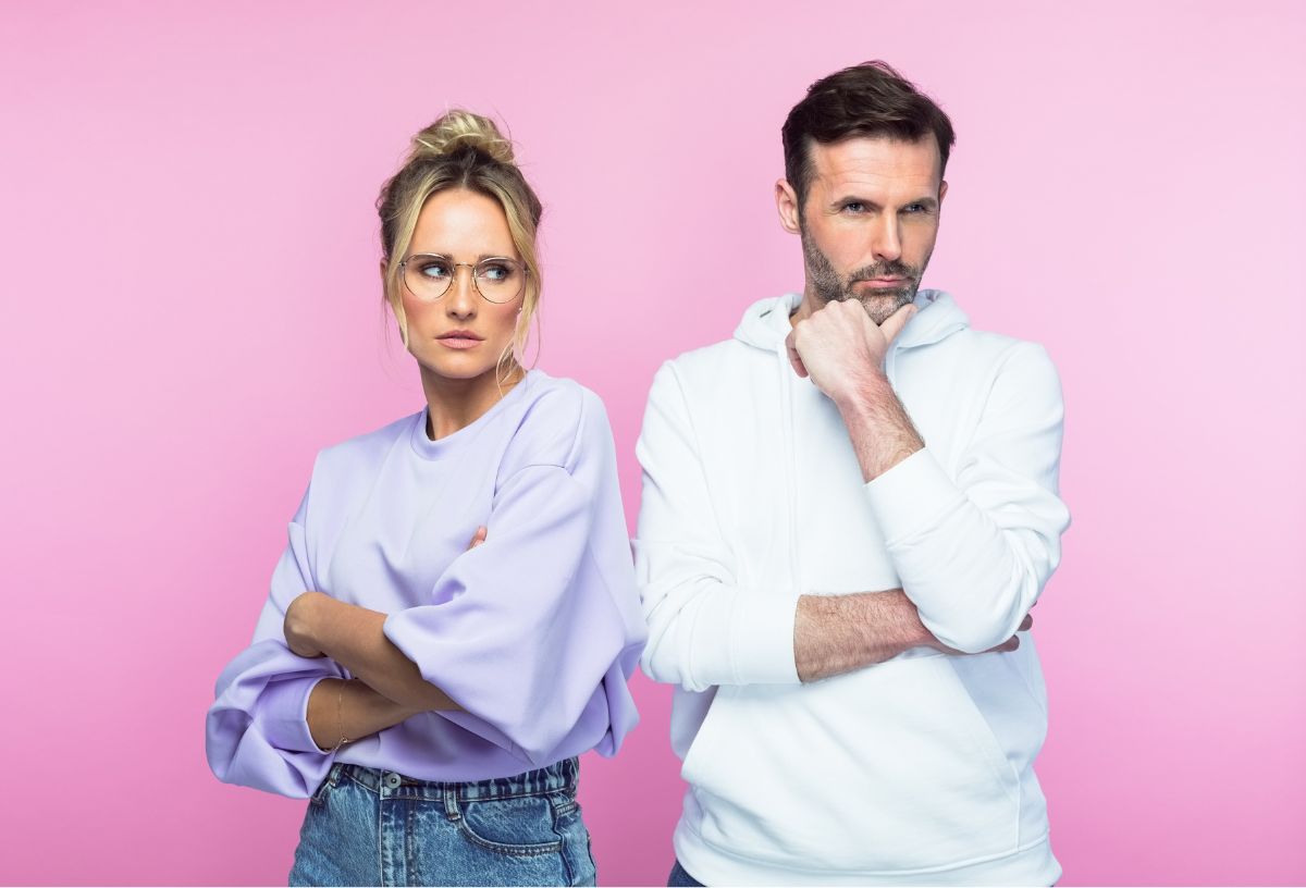 Szőke nő felkontyolt hajjal, szemüvegben, oversized lila felsőben, farmerben, karbatett kézzel bosszúsan néz fehér pulcsis, szakállas férfira, rózsaszín háttér