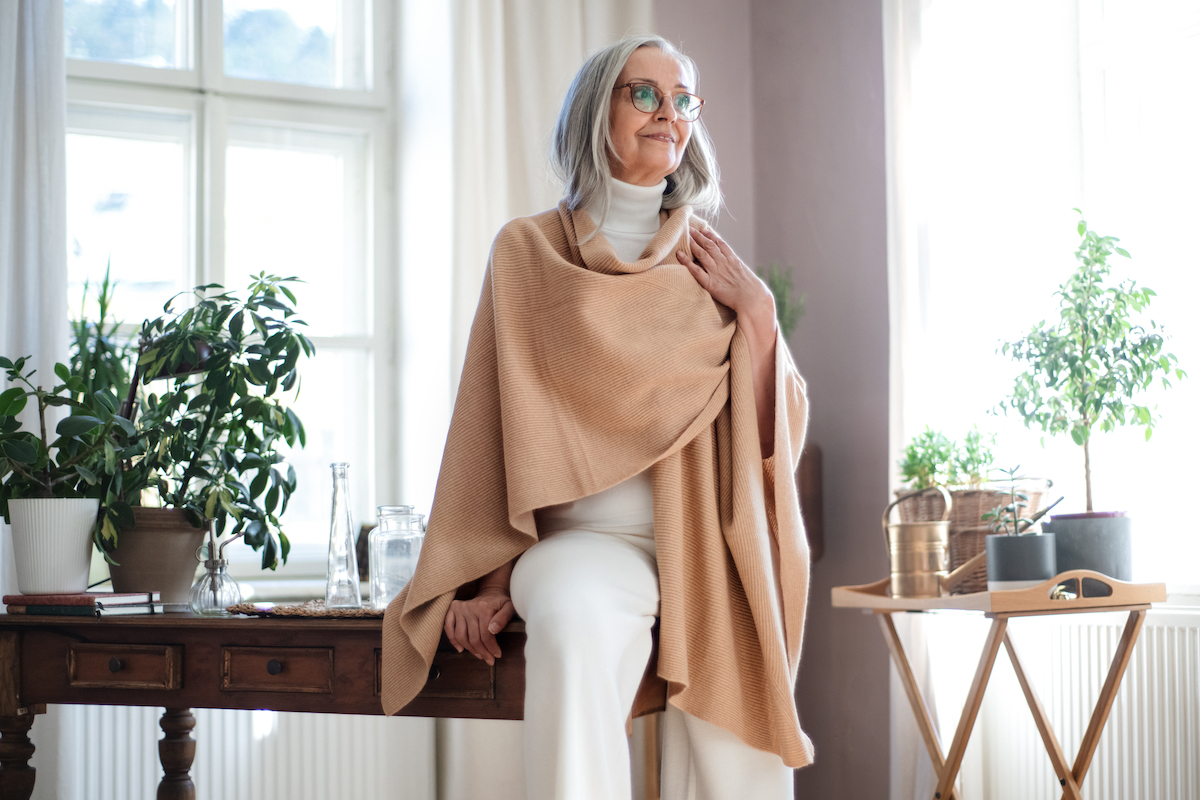 idősebb, fehérbe és bézs kendőbe öltözött szemüveges nő íróasztalra támaszkodik