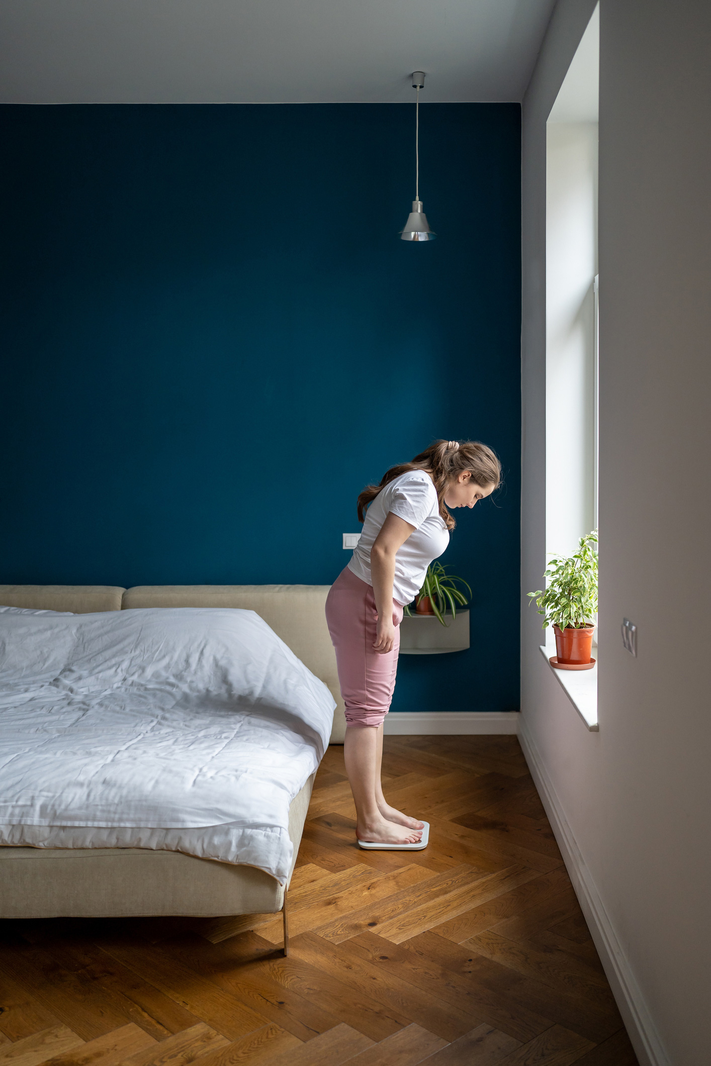 Egy teltebb nő mérlegen áll a hálószobában az ágy mellett.