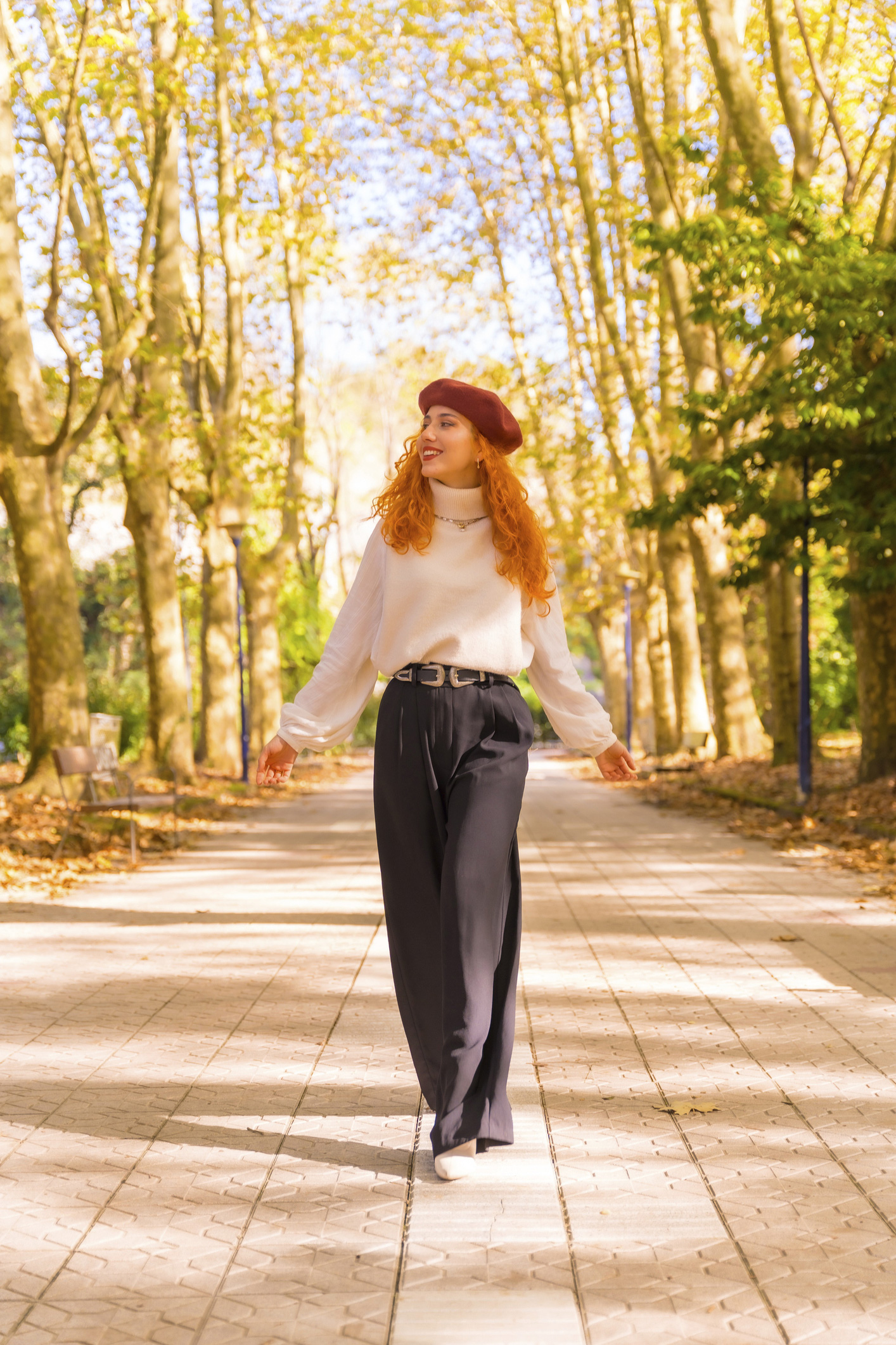 Vörös hajú nő sétál egy városi parkban mosolyogva a napsütésben