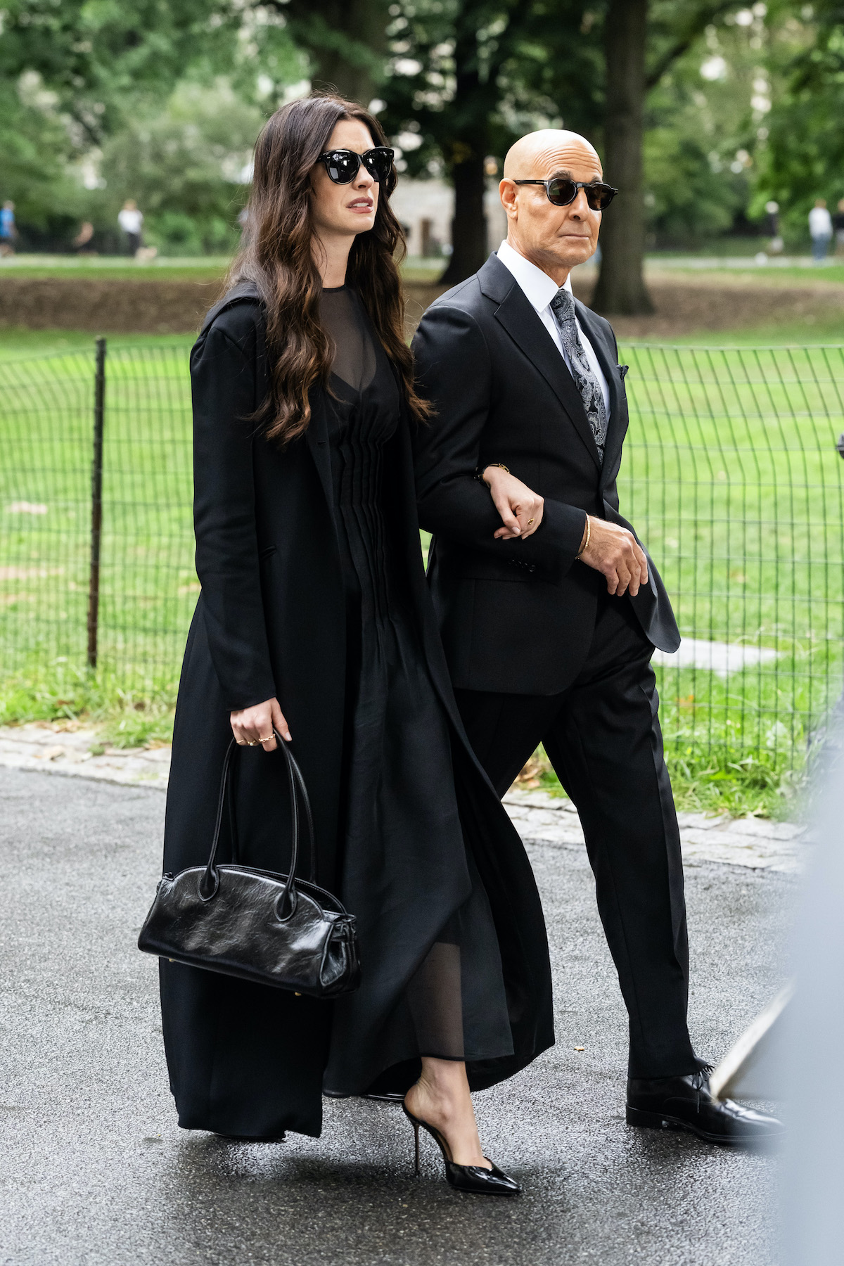 Anne Hathaway és Stanley Tucci fekete ruhában sétál az utcán, a háttérben zöld fű