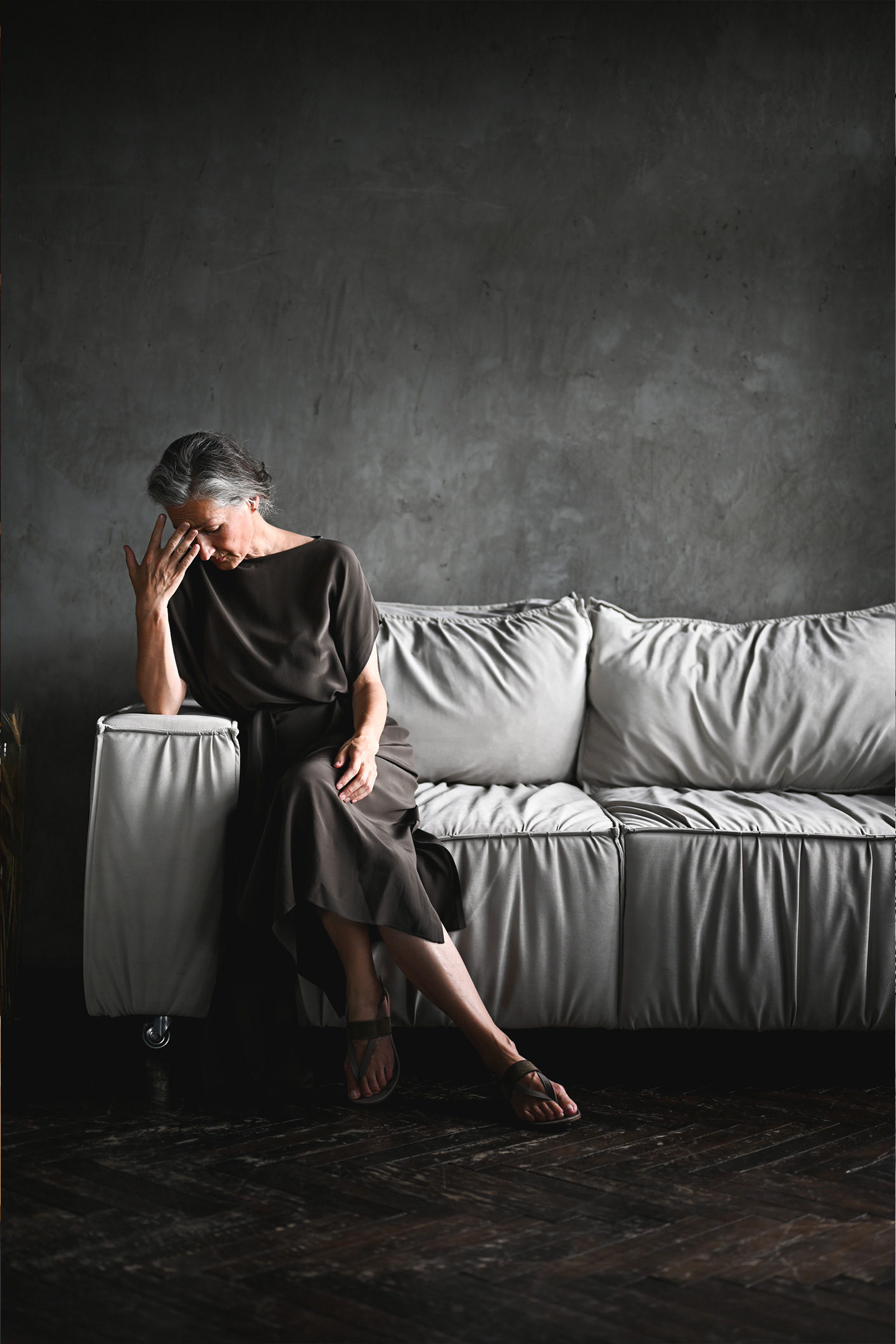 Egy idősebb nő ül összetörve a világos kanapén a sötét szobában