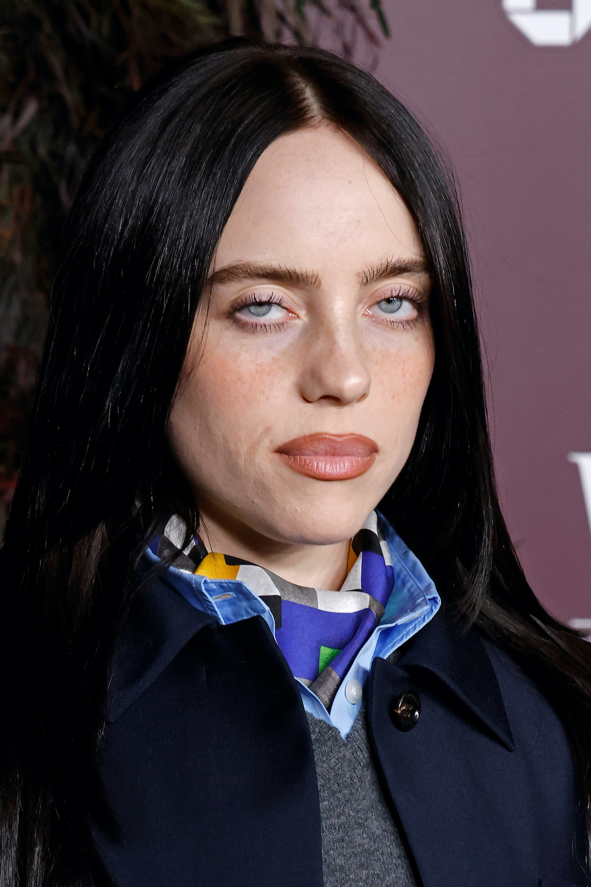 Billie Eilish színes kendővel a nyakán, lila háttér előtt