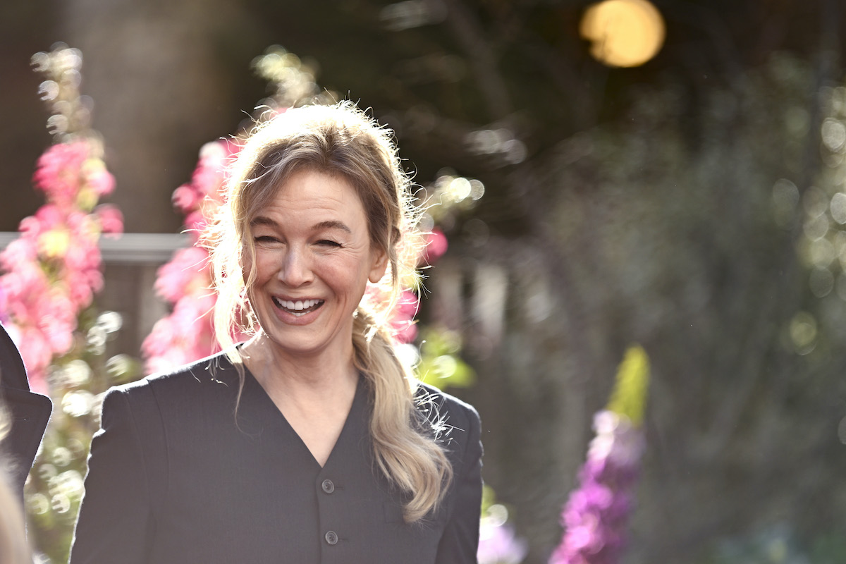 Renée Zellweger virágos háttér előtt, szőke copffal nevet