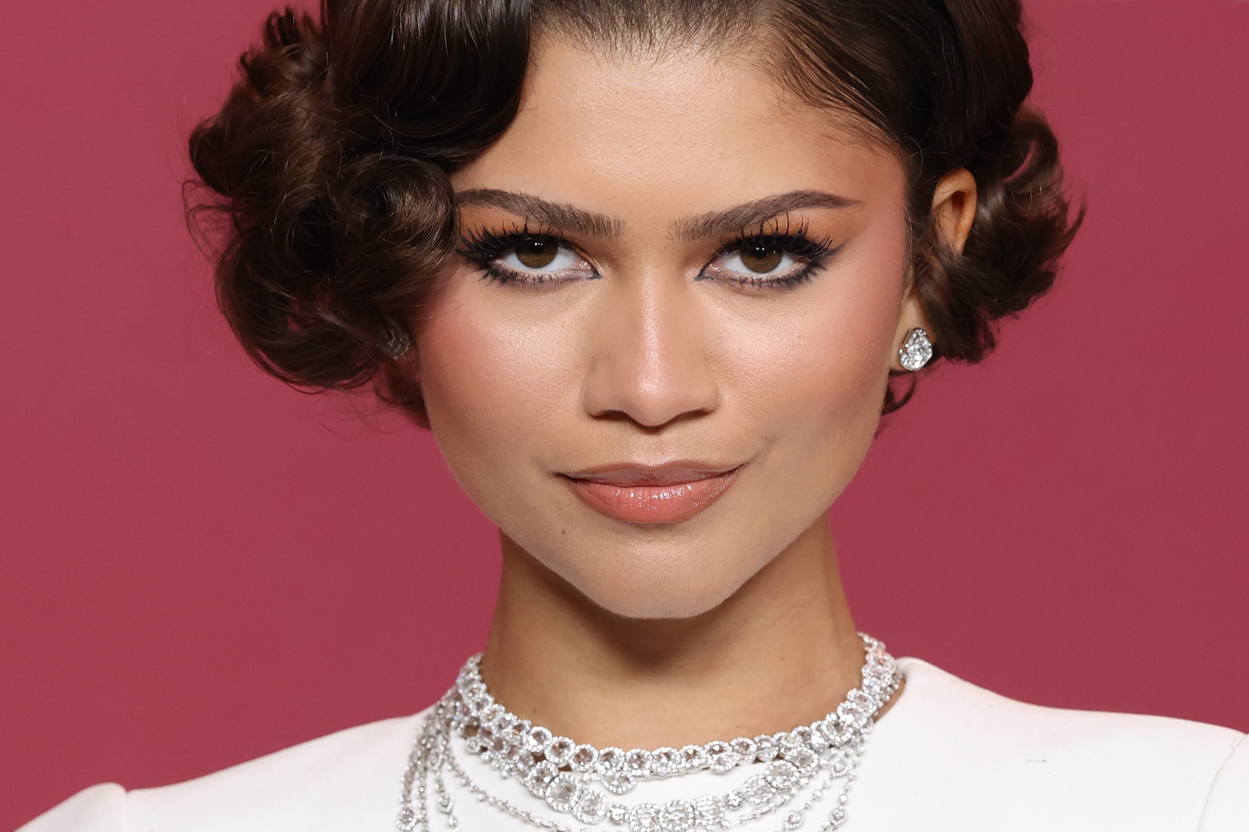 Zendaya közeli felvételen, bordó háttér előtt, fehér felsőben, ékszerekkel