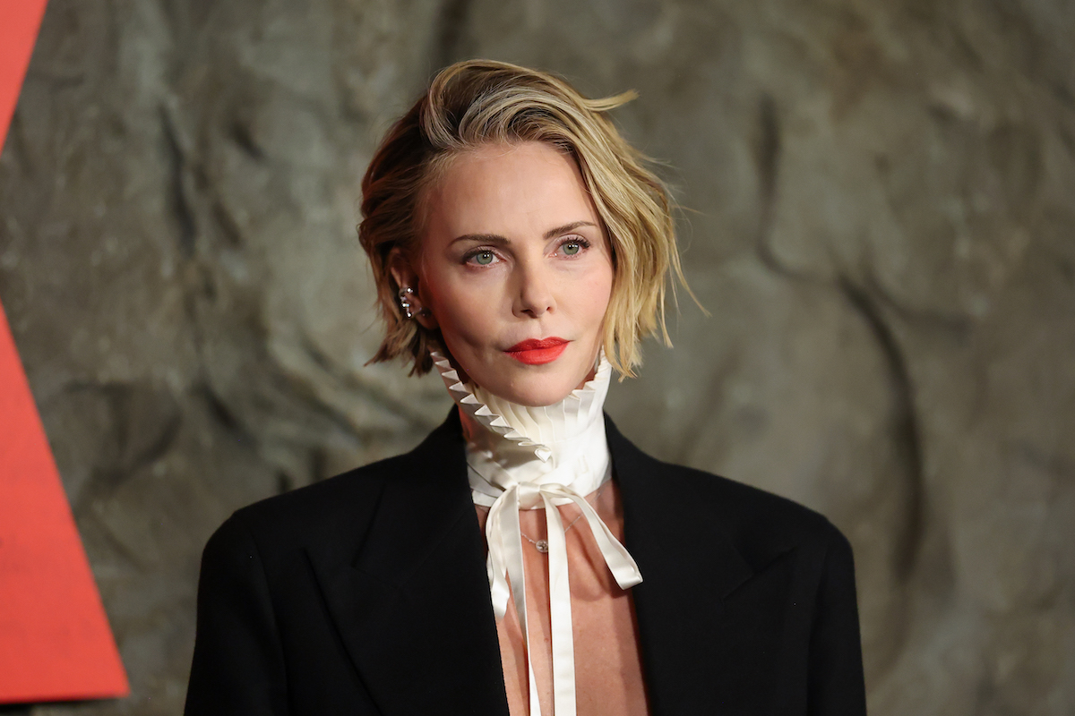 Charlize Theron fehér fodros gallérral és fekete kosztümben, mely alatt nincs semmi – mellvonal felett elvágott fotó