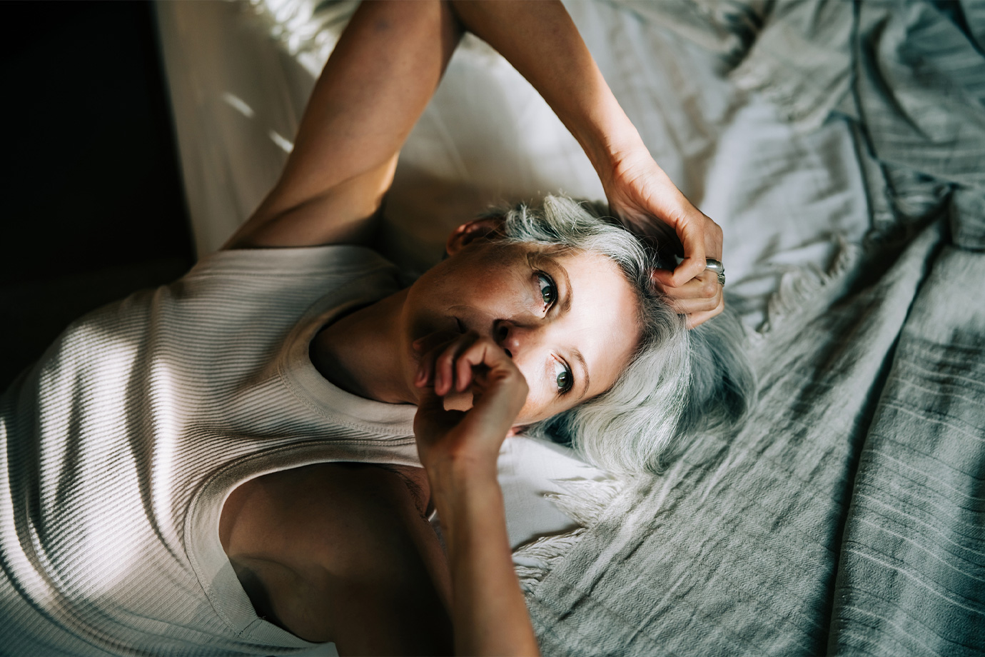 Egy középkorú, ősz hajú nő fekszik hanyatt az ágyban, erős napfény világítja meg az árnyékban