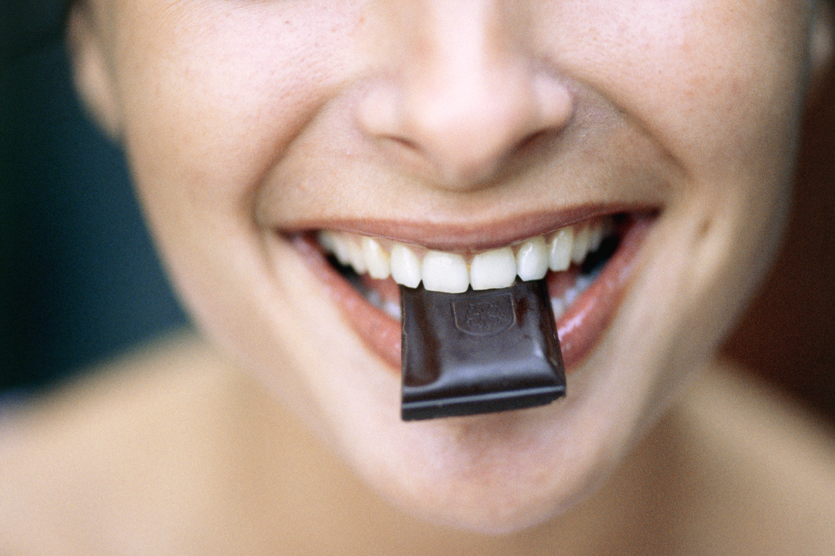 Közeli kép egy nő fogsoráról, amint egy kocka étcsokoládét szorít közé