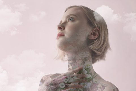 Egy nő, akinek testét virágok borítják, mintha virág lelke lenne, néz a magasba, és a háttérben rózsaszín ég, felhők veszik körbe