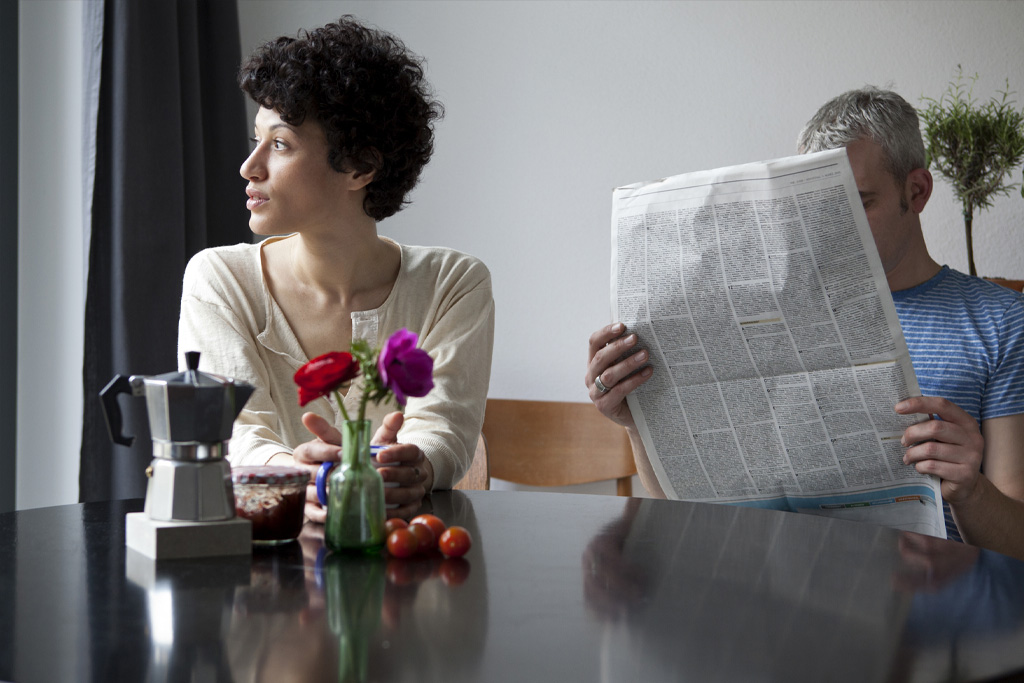 Egy pár ül az asztalnál, a nő szomorúan néz oldalra, míg a férfi újságot olvas