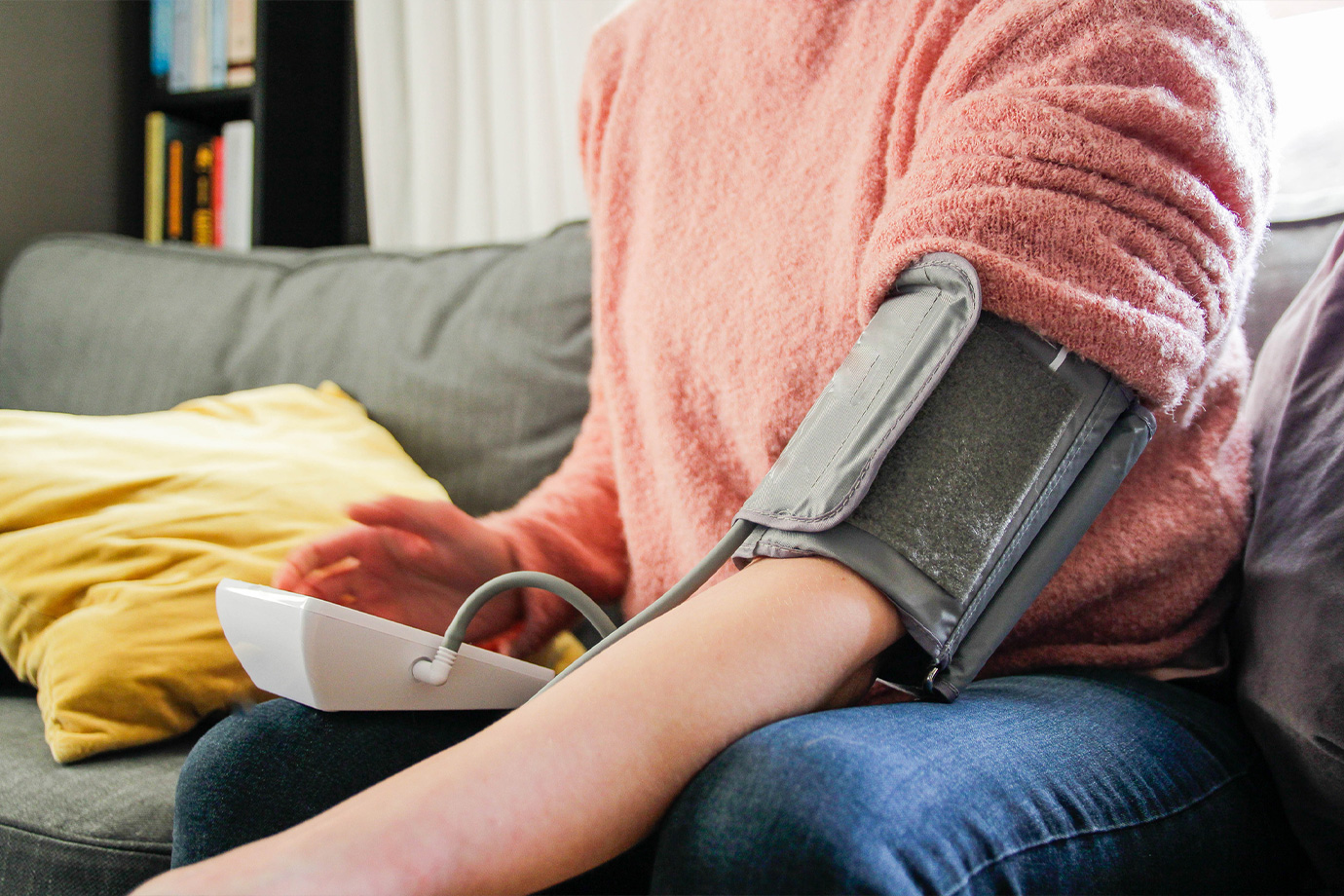 Egy rózsaszín pulóveres nő méri a vérnyomását a nappaliban.