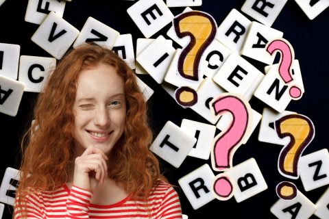 Fekete háttéren Scrabble-betűk, vörös hajú lány kacsint piros-fehér csíkos pólóban, sárga és rózsaszín kérdőjelek