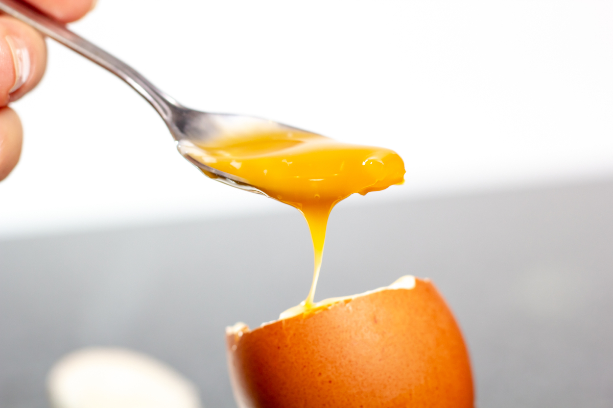 Lágytojás világos háttér előtt, amelyből a sárgáját kanalazza ki egy kéz