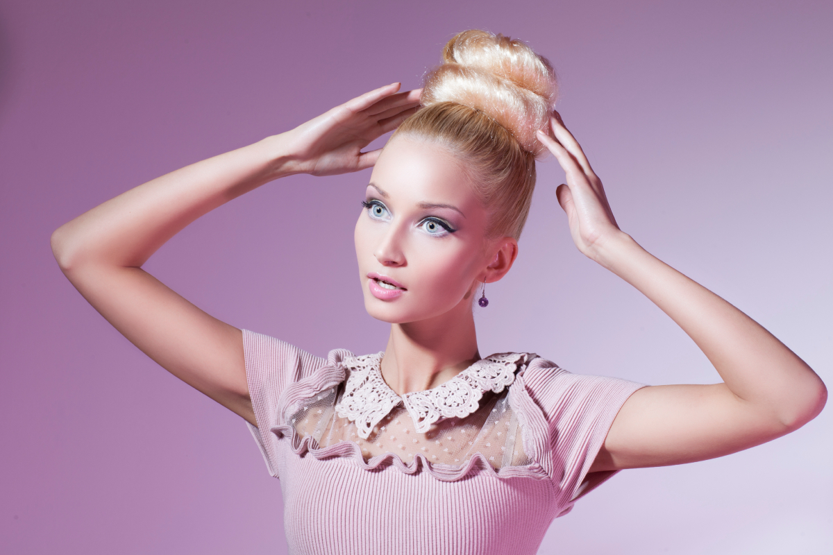Szőke hajú nő rózsaszín háttér előtt rózsaszín ruhában, úgy pózól, mint egy Barbie-baba