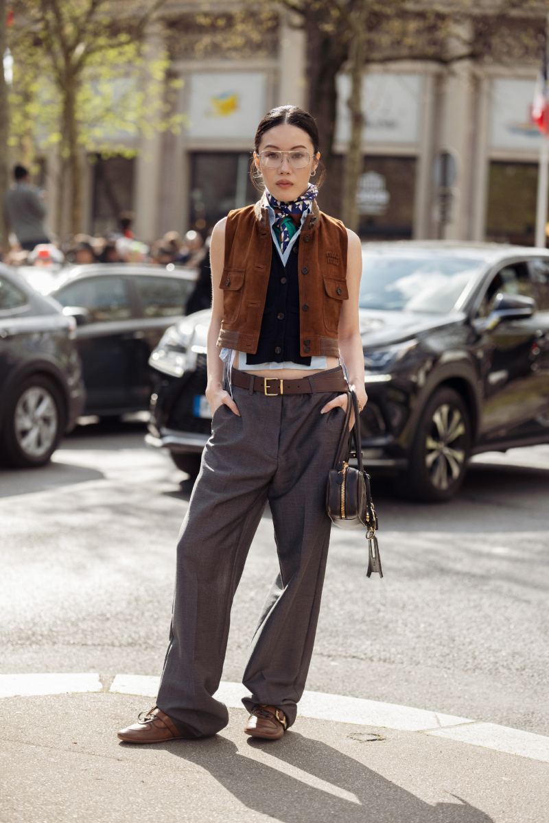 Jenny Tsang barna velúrbőr ujjatlan mellényt visel egy sötét színű, gombos mellény és egy fehér ing fölött, mintás nyaksállal és átlátszó keretes szemüveggel kiegészítve, szürke bő szárú nadrággal párosítva, kis fekete válltáskát hord, és barna bőr loaferben érkezik a Miu Miu show-ra