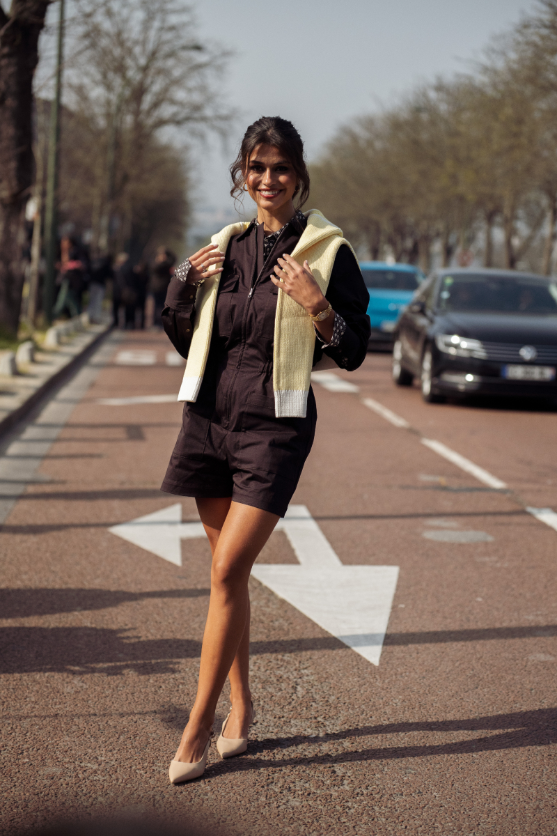 Egy vendég sötétbarna, hosszú ujjú, rövid playsuitot visel elöl zsebekkel és cipzáros részlettel, krémszínű pulóvert vetve a vállára, karkötőkkel és órával, valamint bézs hegyes orrú magas sarkú cipőben jelenik meg a Lacoste show-ra