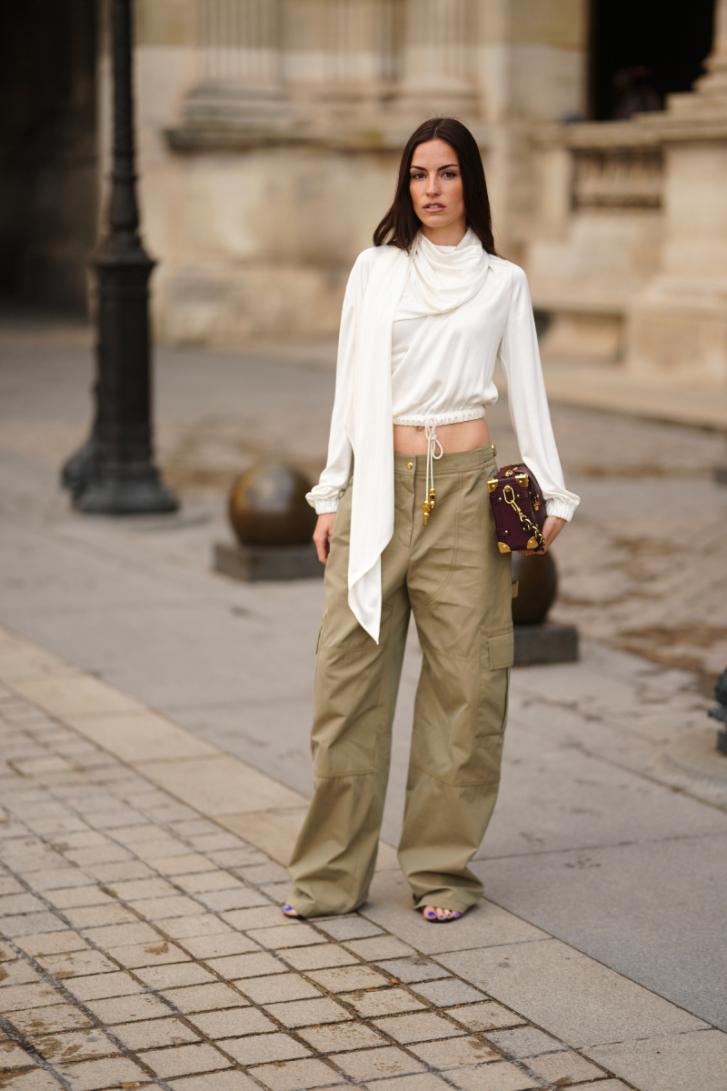 Egy barna hajú nő fehér, hosszú ujjú, drapírozott selyemblúzt visel zsinóros aljjal és sál részlettel, khaki bő szárú cargo nadrágot arany fémdíszekkel, fekete nyitott orrú szandált kék körömlakkal, miközben egy bordó dobozkuplungot hord arany lánccal és sarokmegerősítésekkel