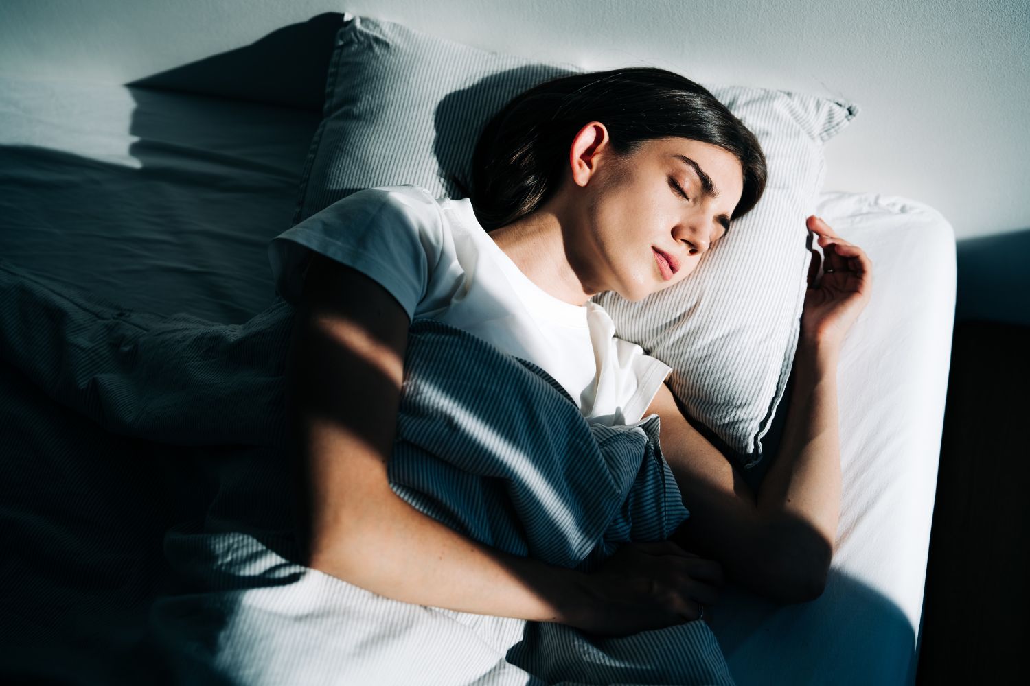 Fiatal, sötét hajú nő kényelmesen alszik fehér ágyneműben, árnyék és fény vetül rá