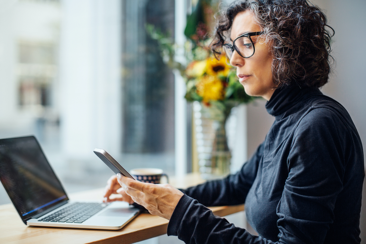 középkorú, göndör, őszülő hajú nő a laptopja előtt ül, kezében mobiltelefon, a kijelzőt nézi