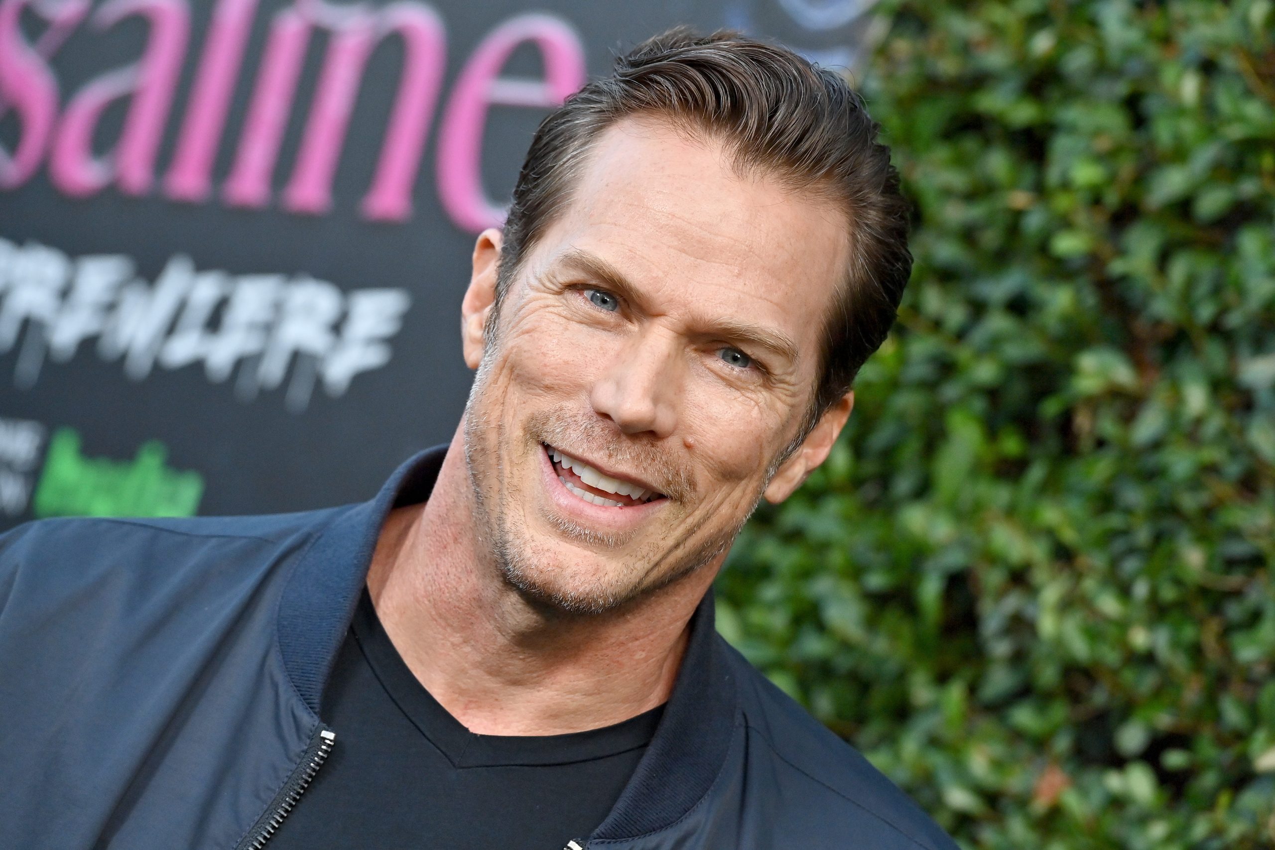 Jason Lewis fekete pólóban és dzsekiben, zöld háttér előtt egy premieren