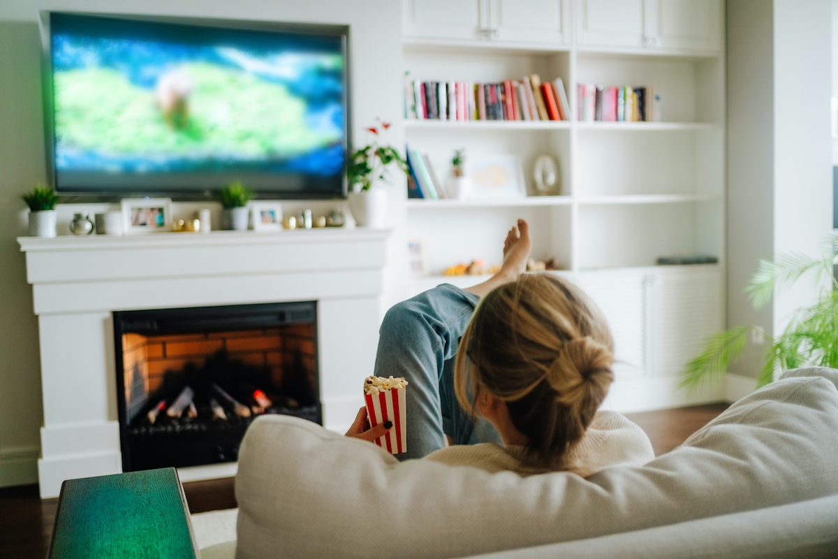 szőke nő háttal, a kezében pattogatott kukoricával tévét néz, a tévé alatt kandalló